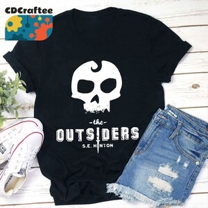 May include: Black t-shirt with a white skull graphic and the text "The Outsiders" with the author's name "S.E. Hinton" underneath.