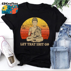 May include: Black t-shirt with a vintage-style illustration of a meditating Buddha in front of a sunset. The text "Let That Shit Go" is printed below the image.