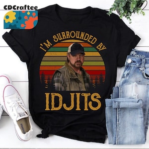 Può includere: T-shirt nero con grafica di un uomo con berretto e gilet, su sfondo tramonto. Il testo "I'M SURROUNDED BY IDJITS" è stampato in stile vintage. Sono visibili scarpe da ginnastica bianche e jeans blu.