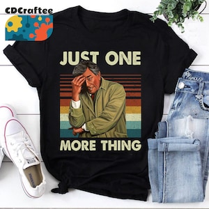 May include: A black t-shirt featuring a vintage-style graphic of a man with a worried expression. The graphic includes the text "Just One More Thing" in white letters.