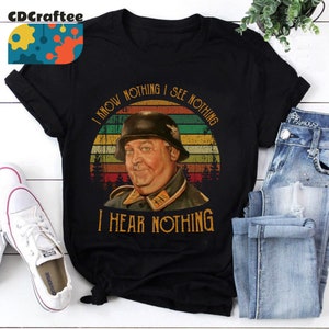 May include: Black t-shirt with a vintage-style graphic featuring a man in a military helmet and uniform, set against a sunset backdrop. The text reads "I KNOW NOTHING I SEE NOTHING I HEAR NOTHING".