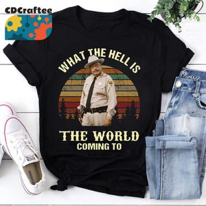 May include: A black t-shirt with a vintage-style graphic of a man in a sheriff's uniform standing in front of a sunset. The text "What the hell is the world coming to" is printed on the shirt.