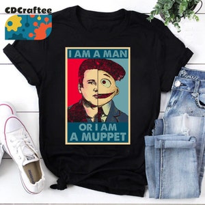 May include: A black t-shirt with a vintage-style poster design featuring a split image of a man and a Muppet. The poster text reads "I am a man or I am a Muppet".