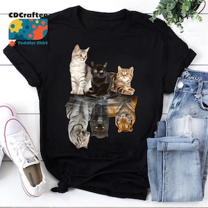 May include: Black t-shirt with a graphic of four cats reflected in water as tigers. The cats are a tabby, a black cat, and two calico cats. The tigers are reflected upside down.