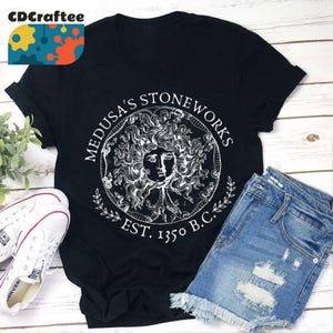 May include: Black t-shirt with a white graphic of Medusa's Stoneworks, a circular design with a stylized image of Medusa's head and the text "Medusa's Stoneworks Est. 1350 B.C."
