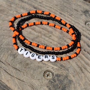 May include: A layered beaded necklace with orange and black beads. The necklace has white letter beads that spell out the word "TIGERS".