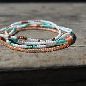May include: A set of four beaded bracelets in white, peach, and turquoise colors. The bracelets are stacked on top of each other, creating a layered look.