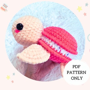Puede incluir: Un juguete de tortuga amigurumi tejido a crochet con una cáscara de macaron rosa y blanca. La tortuga tiene una nariz rosa y ojos negros. El texto "PDF PATTERN ONLY" es visible en la imagen.