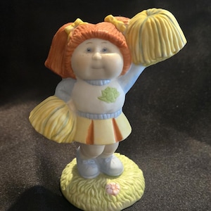Könnte beinhalten: Eine Keramikfigur einer Cheerleaderin mit orangefarbenen Zöpfen, bekleidet mit einem weißen Pullover mit grünem Emblem, einem gelb-orangen Faltenrock und hellblauen Schuhen. Die Figur hält zwei gelbe Pompons und steht auf einem grünen Sockel.