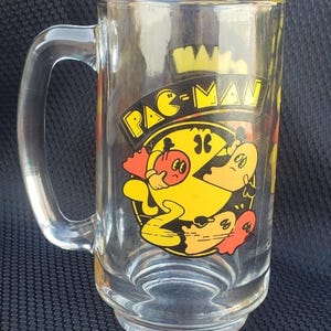 May include: A clear glass Pac-Man mug with a yellow Pac-Man character chasing a red ghost. The mug has a black handle and the words "Pac-Man" printed on the front.