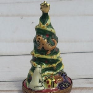 以下が含まれることがあります： クリスマスツリーの形をした小さな円錐形の磁器製小物入れ。ツリーは緑色で、黄色のガーランド、金色の星のトッパー、茶色の猫、白い犬、そして土台にはプレゼントが描かれています。赤い円形の土台と金色の金属製ヒンジが付いています。