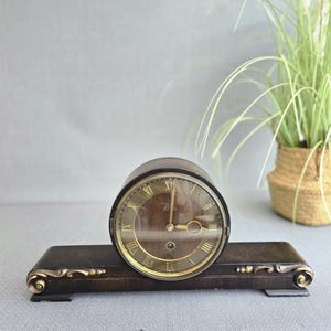 May include: A vintage wooden clock with a round face and Roman numerals. The clock has a dark brown base with gold accents and a key lying in front. The clock face is brown with gold hands and a gold rim.
