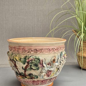 May include: A ceramic planter featuring a cream-coloured base with embossed relief details. The design includes painted scenes of cherubs and trees in shades of green, pink, and brown, bordered by decorative bands of laurel leaves and foliage.