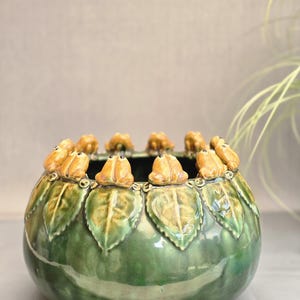 May include: A round ceramic planter with a glossy dark green glaze featuring a relief pattern of overlapping leaves. The rim is decorated with a series of small, light brown sculpted frogs positioned around the top edge of the vessel.