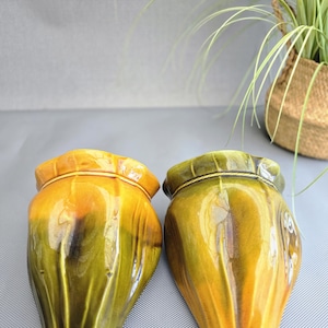 May include: Two ceramic vases with a glossy finish. The vases are teardrop-shaped, with a blend of yellow and green hues. The tops have a decorative rim. A woven basket with greenery is in the background.