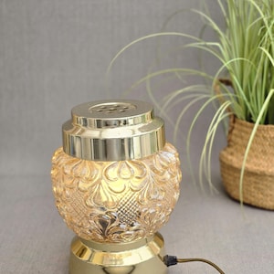 May include: A table lamp featuring a textured, amber-coloured glass shade with a repeating floral pattern. The lamp has a polished gold-tone metal base and a matching gold-tone metal top cap with a series of small circular ventilation holes.