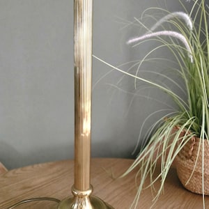 May include: A brass table lamp with a fluted column and a round base. The lamp has a standard bulb socket and a cord. The lamp is on a wooden table. The lamp is approximately 38 cm tall.