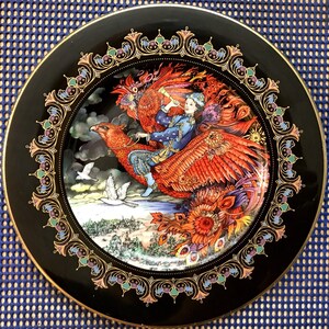 May include: A black ceramic plate with a gold rim and a colorful image of a person riding a phoenix. The image is surrounded by a decorative border.