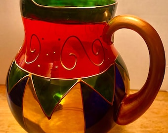 May include: A glass pitcher featuring a red upper section with gold swirl and dot accents, a green rim, and a lower body decorated with alternating triangular panels of green, blue, and gold. The pitcher includes a solid gold-colored handle.