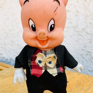 Puede incluir: Un cerdo rosa con un sombrero rojo y un traje negro con un chaleco a cuadros y una corbata de lazo. El cerdo tiene una expresi&oacute;n feliz y est&aacute; de pie sobre una superficie de madera.