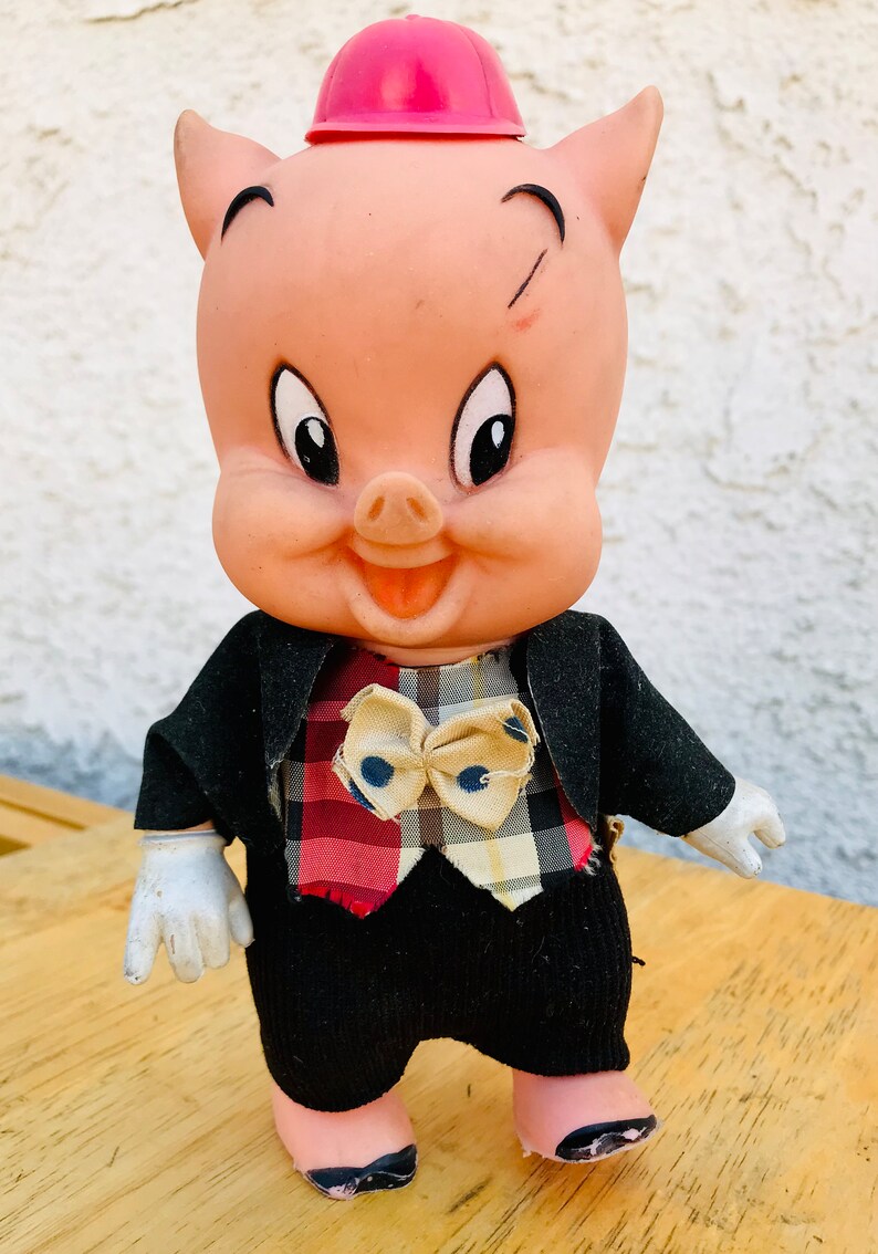 Puede incluir: Un cerdo rosa con un sombrero rojo y un traje negro con un chaleco a cuadros y una corbata de lazo. El cerdo tiene una expresi&oacute;n feliz y est&aacute; de pie sobre una superficie de madera.