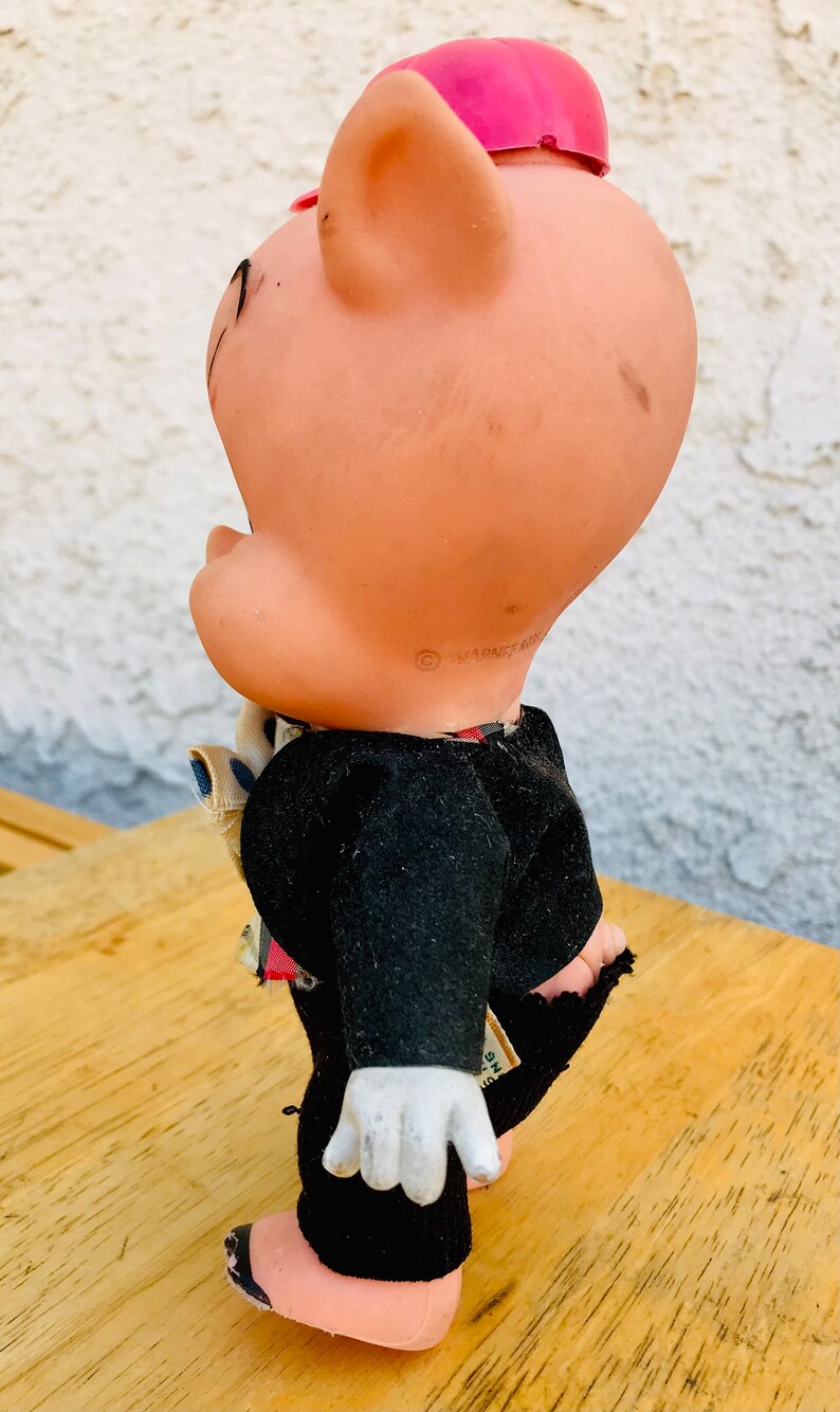 Puede incluir: Un juguete de cerdo vintage que lleva un traje negro y un sombrero rosa. El cerdo tiene una camisa blanca con una corbata de patr&oacute;n azul y blanco. El juguete est&aacute; hecho de pl&aacute;stico y tiene una cara pintada.