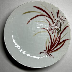May include: White ceramic plate with a red and gold floral design. The design features stylized flowers and leaves.