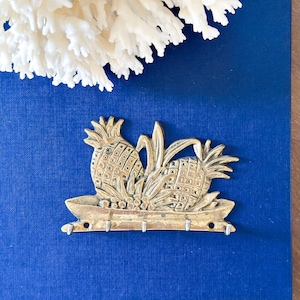 May include: A brass pineapple key holder with three hooks, featuring two detailed pineapples and leaves. The key holder is set against a deep blue background, with white coral in the upper left corner.