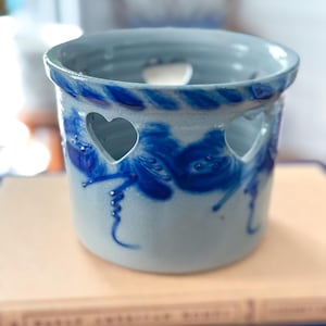 Puede incluir: Un portavelas de cerámica con base azul claro y diseños florales azul oscuro. El portavelas tiene recortes en forma de corazón y un borde decorativo. El portavelas está sobre un libro.