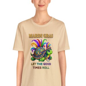 May include: A beige t-shirt featuring a colourful turtle wearing a Mardi Gras crown and feathers. The text "Mardi Gras" is above the turtle and "Let the good times roll" is below the turtle.