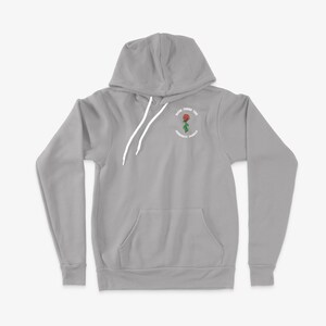 May include: A grey hooded sweatshirt with a white drawstring and a pocket on the front. The sweatshirt has a white embroidered design of a red rose with green leaves and the text "Rise from the Concrete Jungle".