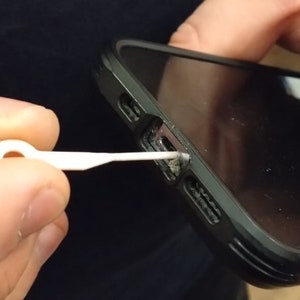 May include: A white plastic tool with a loop at one end is being used to clean out the charging port of a black smartphone.
