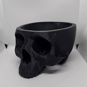 May include: A black skull-shaped planter. The planter is made of a smooth, matte black plastic and has a round opening at the top. The skull has a detailed design with visible eye sockets, a nose, and teeth.