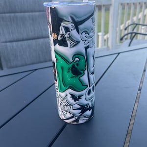 May include: A stainless steel tumbler with a wraparound design featuring characters from The Nightmare Before Christmas. The design includes Jack Skellington, Sally, and other spooky elements in black, white, and green. A white straw is inserted in the lid.