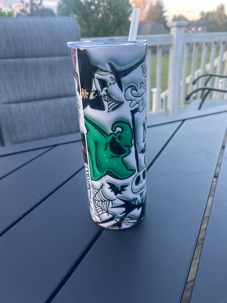 May include: A stainless steel tumbler with a wraparound design featuring characters from The Nightmare Before Christmas. The design includes Jack Skellington, Sally, and other spooky elements in black, white, and green. A white straw is inserted in the lid.