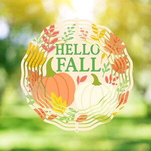 Op de afbeelding: Een cirkelvormige herfstdecoratie met de tekst "HELLO FALL" in het groen. Het ontwerp bevat een oranje en een witte pompoen, omringd door herfstbladeren in rood, geel en groen. De achtergrond is een wazig groen.