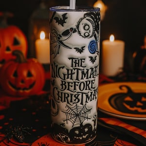 May include: A white tumbler with a straw featuring a design from "The Nightmare Before Christmas." The design includes bats, Jack Skellington, and a spiderweb. The text "The Nightmare Before Christmas" is printed on the tumbler.