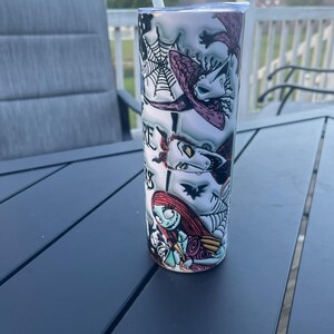 May include: A cylindrical stainless steel tumbler with a design featuring characters from The Nightmare Before Christmas. The design includes images of Sally, Jack Skellington, and other characters, with a white straw and lid. The tumbler sits on a dark grey table.