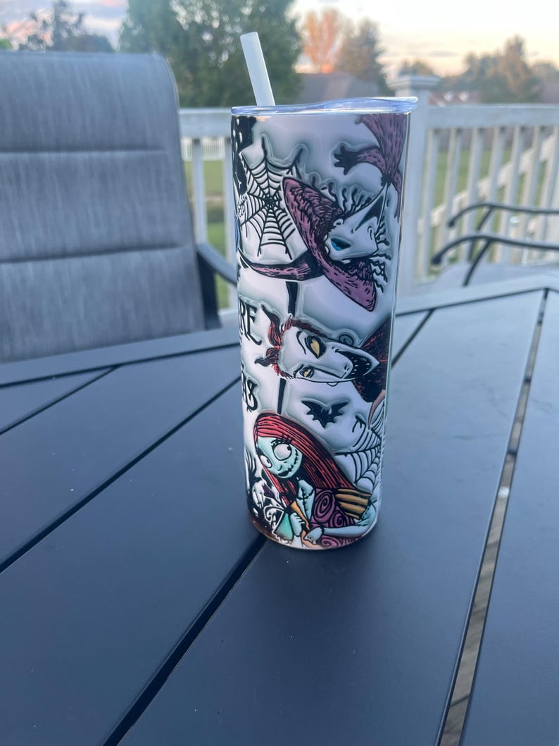 May include: A cylindrical stainless steel tumbler with a design featuring characters from The Nightmare Before Christmas. The design includes images of Sally, Jack Skellington, and other characters, with a white straw and lid. The tumbler sits on a dark grey table.