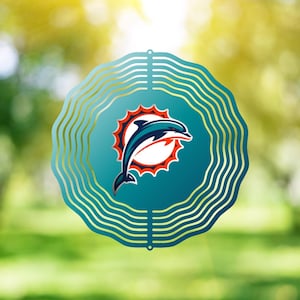 Op de afbeelding: Een teal en gouden metalen windspinner met het Miami Dolphins logo. Het logo bevat een springende dolfijn in teal en wit, tegen een zonnesterontwerp. De spinner heeft een golvend, gelaagd ontwerp.