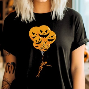 May include: A black t-shirt with a graphic of a skeleton holding onto orange pumpkin-shaped balloons with jack-o'-lantern faces.