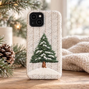 May include: A phone case with a textured, cream-colored knit pattern background. Centered on the back is an embroidered design of a green pine tree with white snow on its branches, standing on a small, white, snow-covered mound.