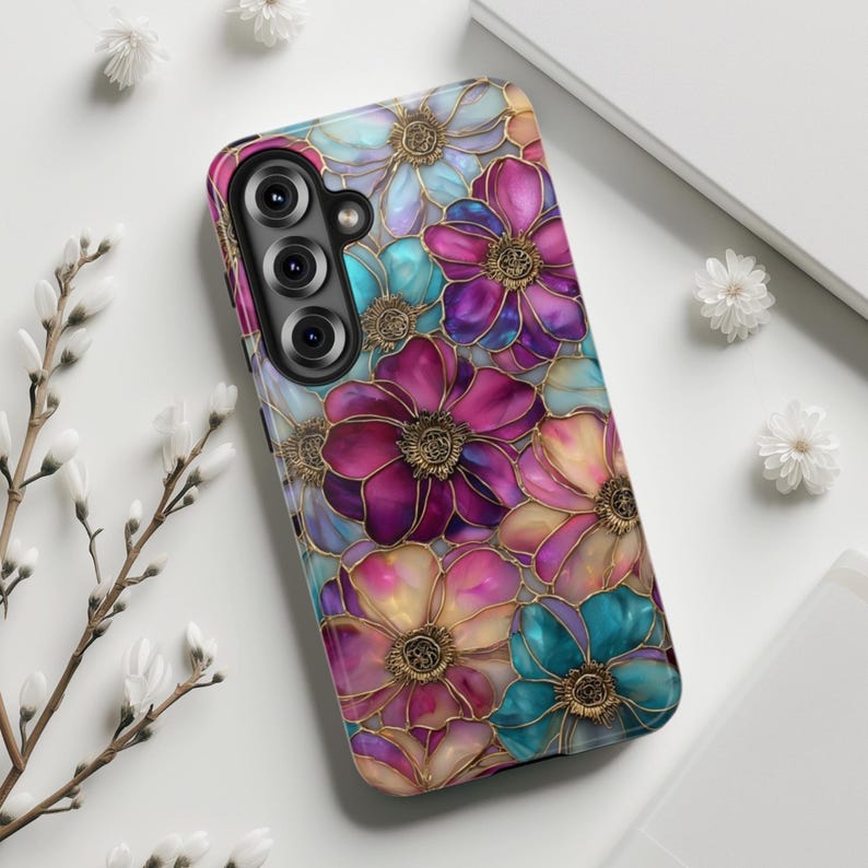 K&ouml;nnte beinhalten: Eine Smartphone-H&uuml;lle mit einem lebendigen Blumenmuster aus &uuml;berlappenden Bl&uuml;tenbl&auml;ttern in Magenta, T&uuml;rkis und sanftem Gelb. Jede Blume ist mit einer metallisch goldenen Kontur versehen, die einen Buntglaseffekt auf der Oberfl&auml;che erzeugt.