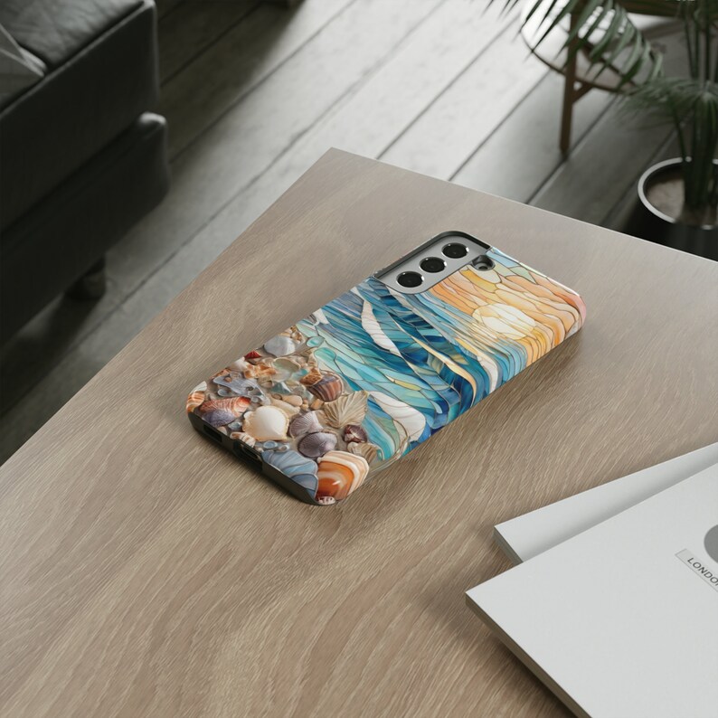 May include: A rectangular smartphone case featuring a printed design of a beach shoreline with scattered seashells, blue ocean waves, and a golden sun. The case has a black frame with cutouts for the camera lenses and flash on the upper back.