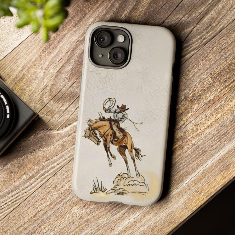 May include: A white iPhone case with a drawing of a cowboy riding a bucking horse. The cowboy is wearing a hat and holding a rope. The horse is brown and white.