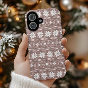 May include: A mobile phone case featuring a repeating Nordic-style pattern of white snowflakes and geometric shapes set against a muted mauve background. The case has a black camera module cutout with two circular lenses positioned at the top left.