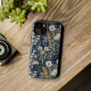 Op de afbeelding: Een blauwe telefoonhoes met een bloemenpatroon met witte madeliefjes, paarse bloemen en groene bladeren. De hoes heeft een uitsparing voor de cameralens en een verhoogde rand rond de randen.