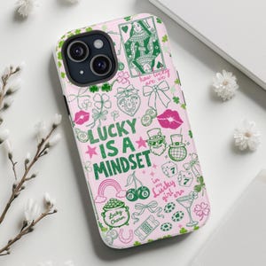 May include: A light pink phone case featuring a green and pink graphic print with the text "LUCKY IS A MINDSET", "how lucky are we", and "in my lucky girl era". The design includes illustrations of a playing card, shamrocks, bows, and dice.