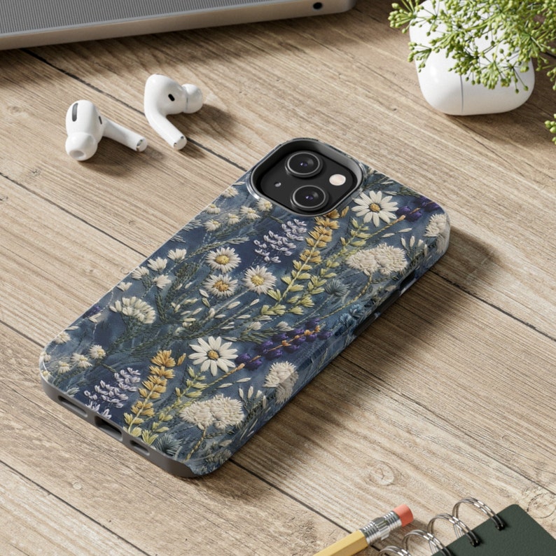 Op de afbeelding: Een blauwe telefoonhoes met een bloemenpatroon met witte bloemen en groene bladeren. De hoes heeft een camera-uitsparing en een verhoogde rand rond de randen.