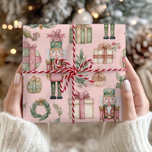May include: A wrapped gift with pink wrapping paper featuring watercolor illustrations of nutcrackers, presents, and greenery. The gift is tied with red and white twine. The background is blurred, suggesting a festive holiday theme.